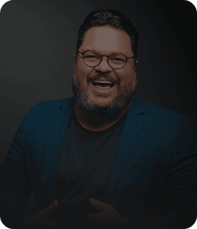 Evento Super Megavendas para concessionárias Um painel digital em cinza e preto com um homem sorridente de óculos, barbudo e camisa social azul. Ele aponta para o centro do painel, onde há vários cartões com textos como 'Equipe engajada', 'Clientes agendados' e 'Meta de vendas batida!'. Na parte inferior, há botões com os textos 'Ligar para cliente', 'Chamar no WhatsApp', 'Agendar tarefa', 'Registrar novo contato' e 'Ver convite'.